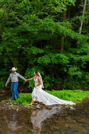 Bride in white gown and groom in cowboy hat hold hands crossing a rocky creek surrounded by lush green forest.