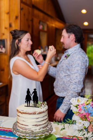 Bride and groom feeding each other wedding cake at rustic barn reception with cowboy silhouette cake topper.