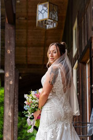 Bride in lace wedding dress and veil holds colorful bouquet outside rustic wooden venue.