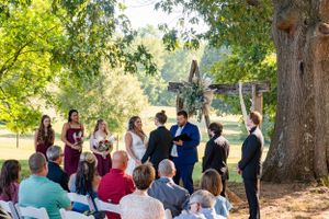 An outdoor wedding ceremony under tall trees with guests seated on white chairs in a natural setting.