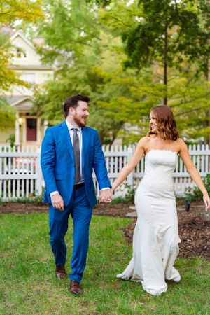 A couple shares a joyful moment while walking on grass in front of a house with a white picket fence.