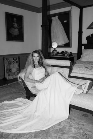 A bride in an elegant white gown poses on an antique chaise lounge in a formal setting.