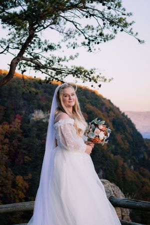 Bride in lace gown poses on mountain overlook during autumn sunset.