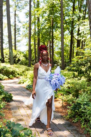 Bride in elegant white dress with thigh slit holds blue hydrangea bouquet while walking on garden path surrounded by trees.