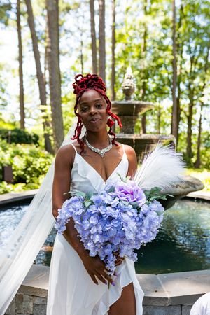 Beautiful bridal portrait by fountain with lavender hydrangea bouquet and white gown in garden setting.
