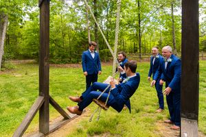 A group in blue suits gathers outdoors around a wooden swing structure on a grassy lawn surrounded by trees.