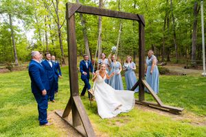 Wedding party gathered around a rustic wooden swing frame in a sunny wooded outdoor setting.