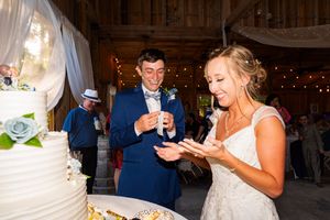Joyful couple feed each other wedding cake during reception in rustic barn venue with string lights and decorations.