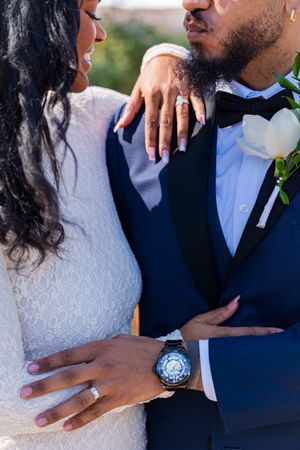 Close-up details of a bride and groom embracing, showing wedding attire and jewelry in an intimate outdoor setting.