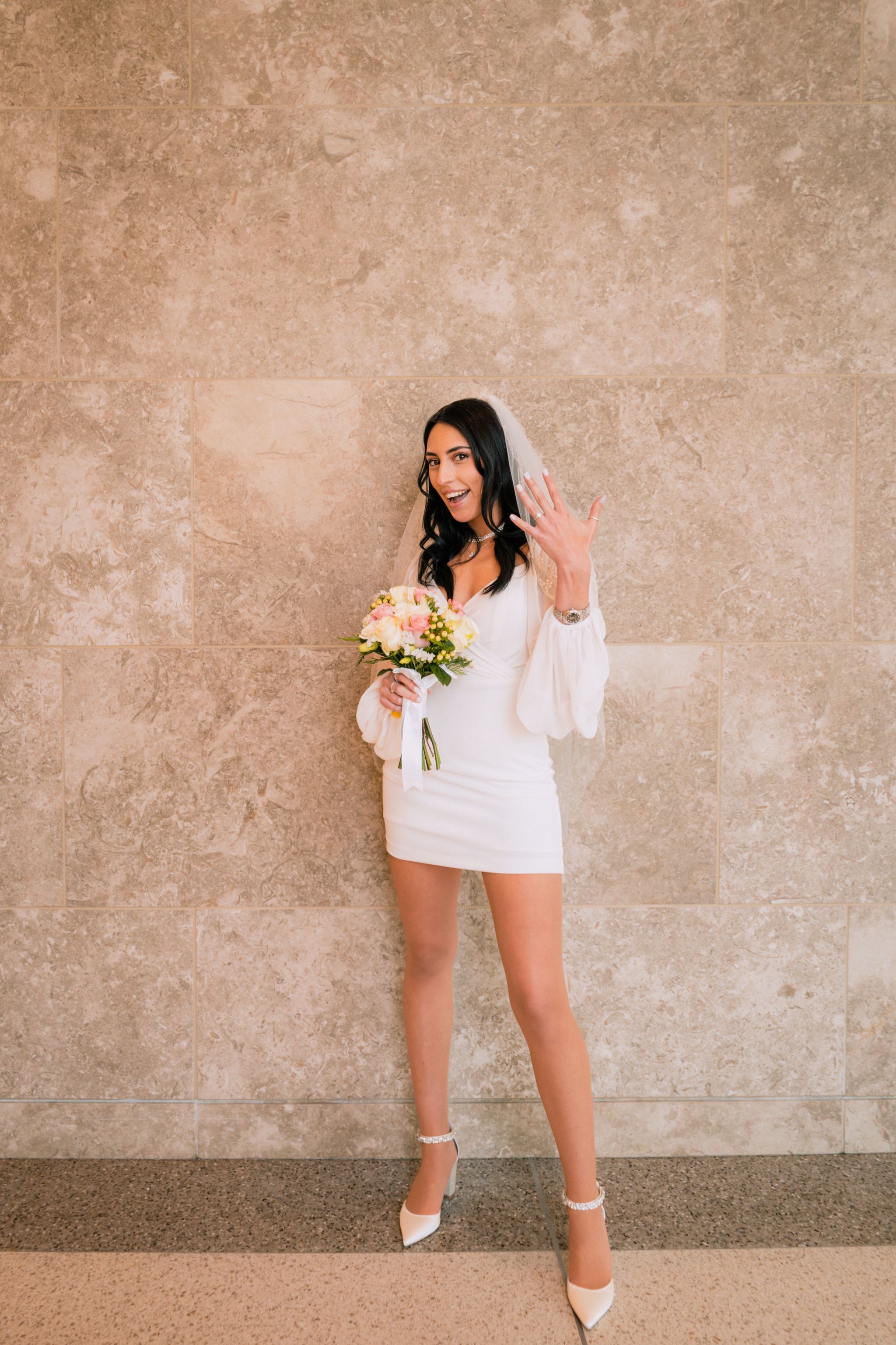 Modern bride in short wedding dress poses against neutral wall with bouquet.