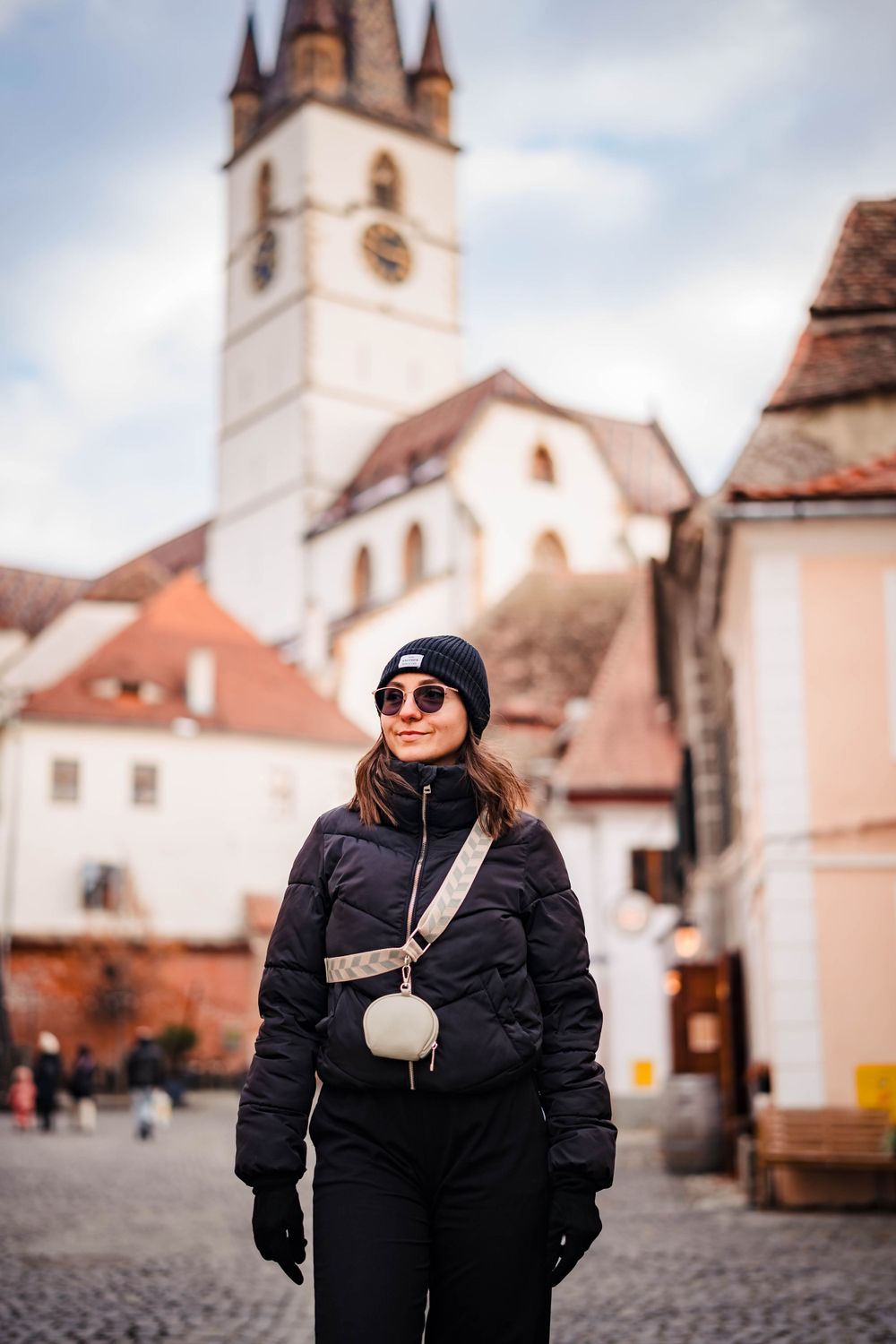 Two people in winter coats pose in front of historic European architecture on a cobblestone square.