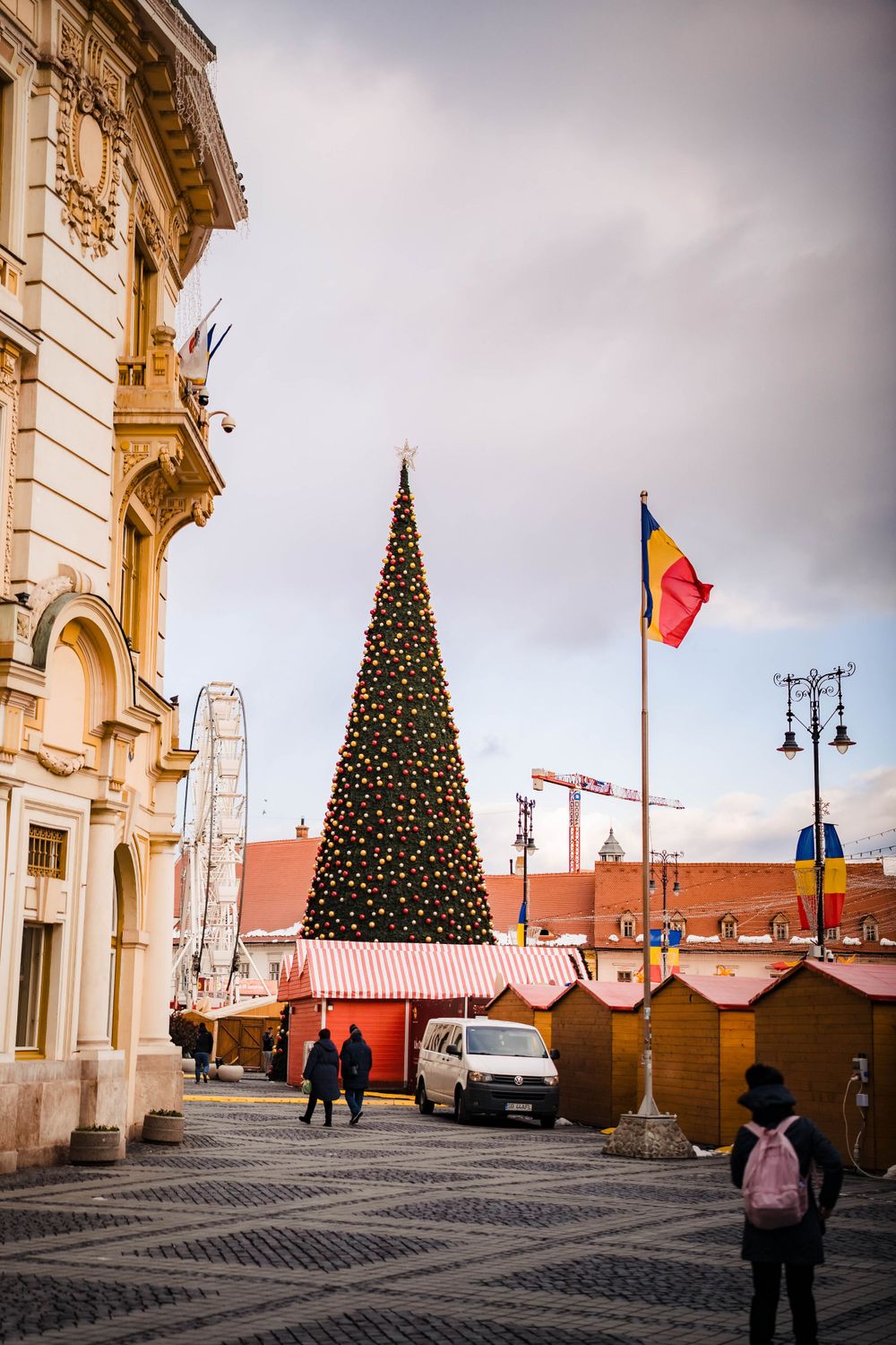 Large Christmas tree and wooden market stalls stand in front of classical architecture.