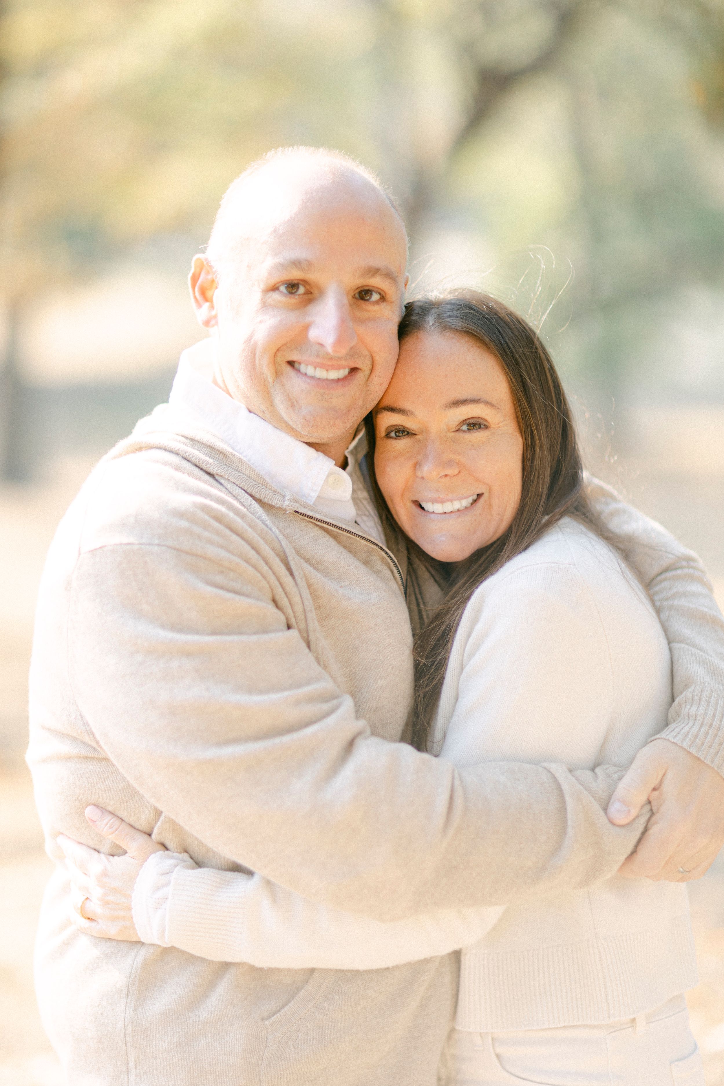 Couple embracing outdoors in cream-colored sweaters with soft natural lighting.