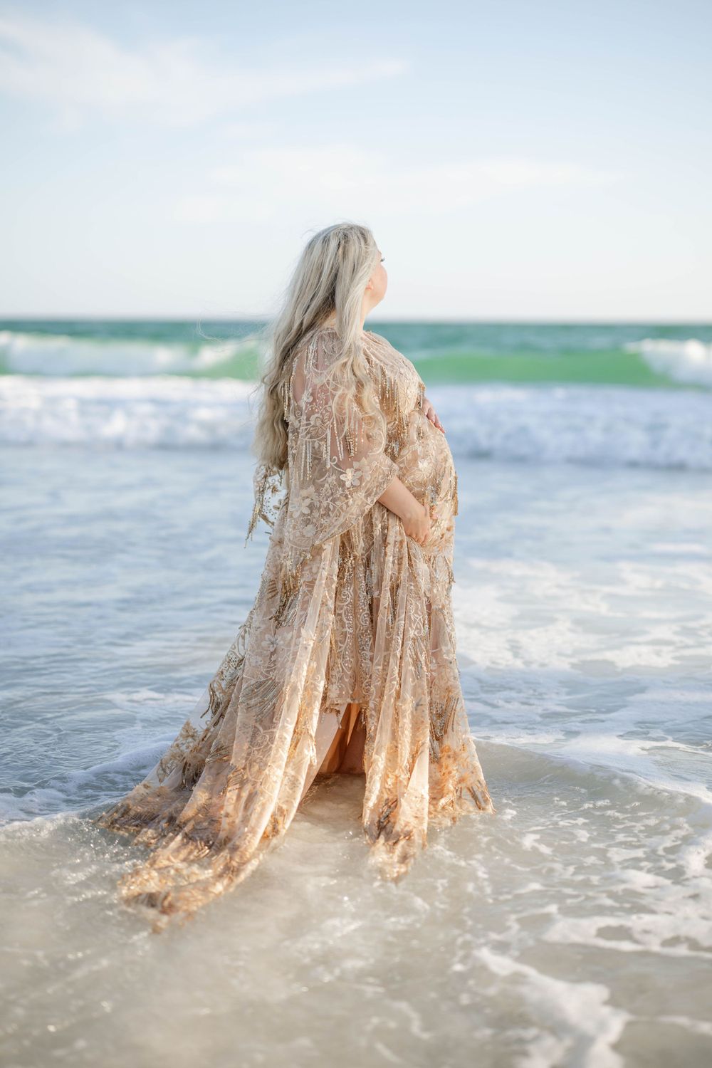 Ethereal beach portrait with lace dress flowing into ocean waves at sunset.