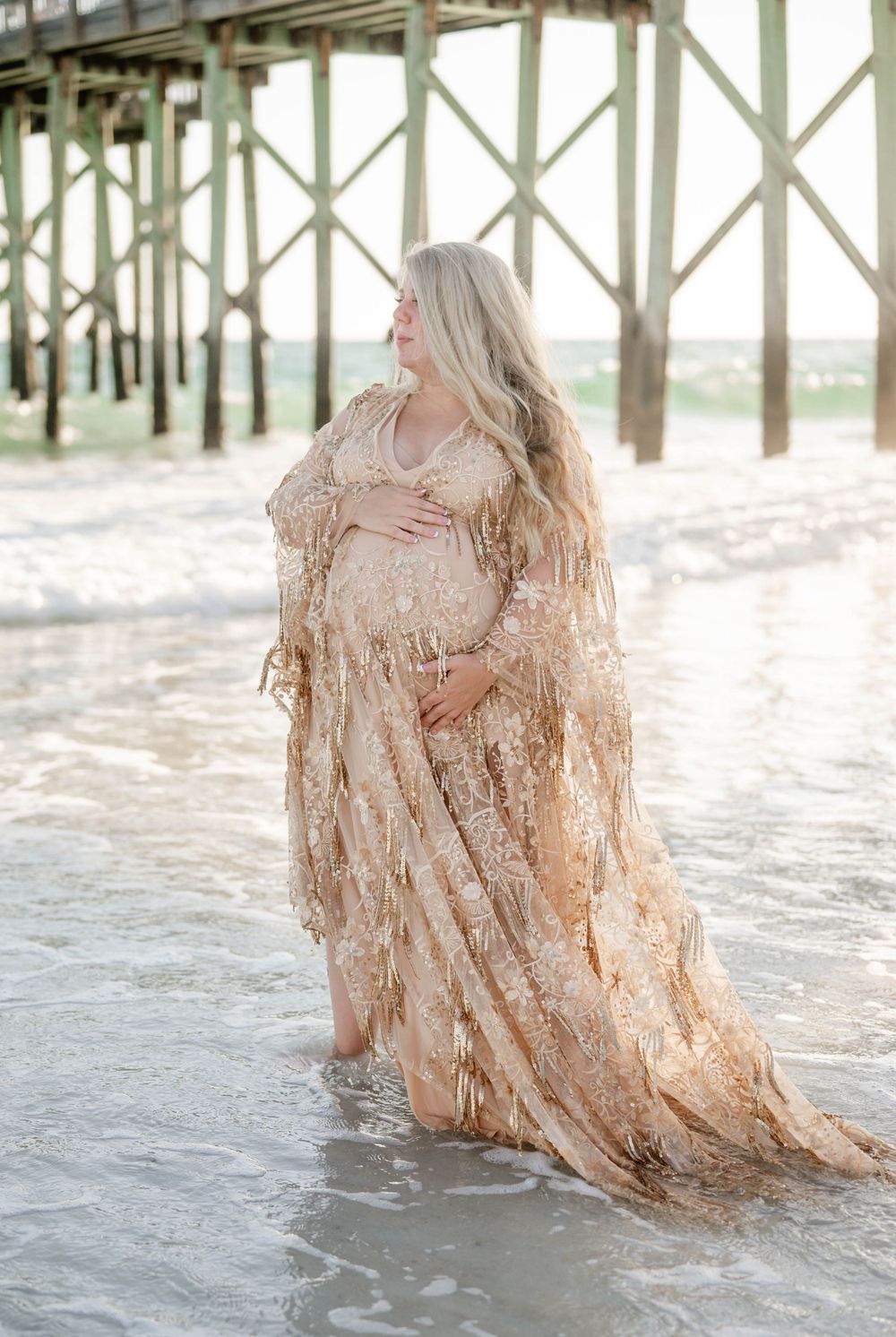 Coastal maternity portrait under pier featuring elegant champagne-colored lace gown.