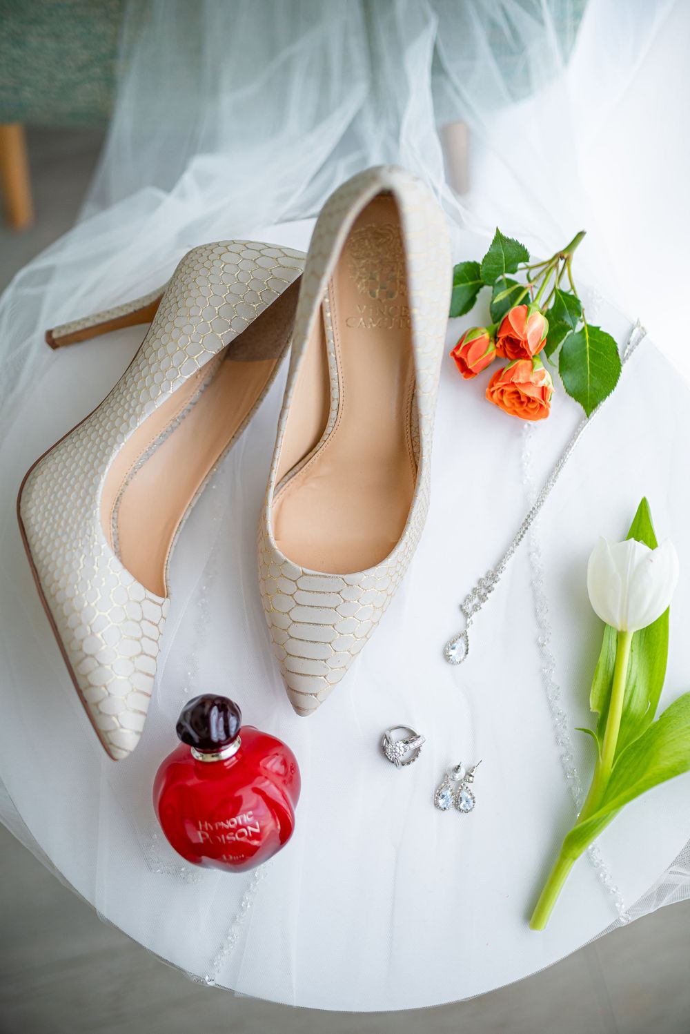 Elegant white bridal heels with red perfume bottle and flowers on tulle fabric.