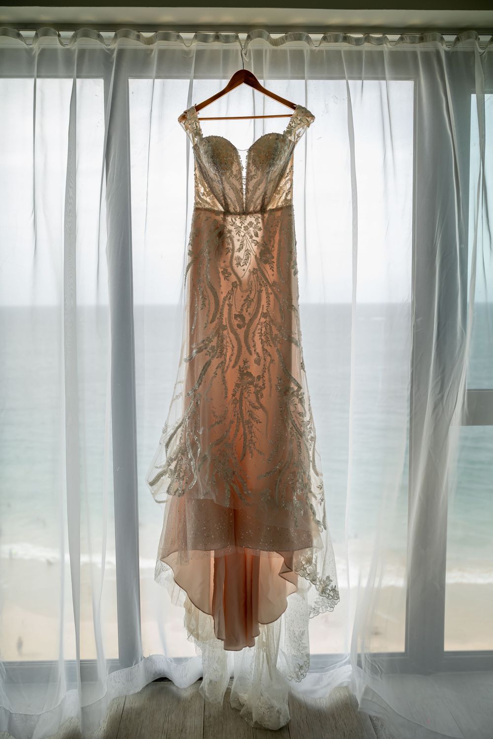 Blush pink lace wedding gown hanging in front of sheer curtains with ocean view.