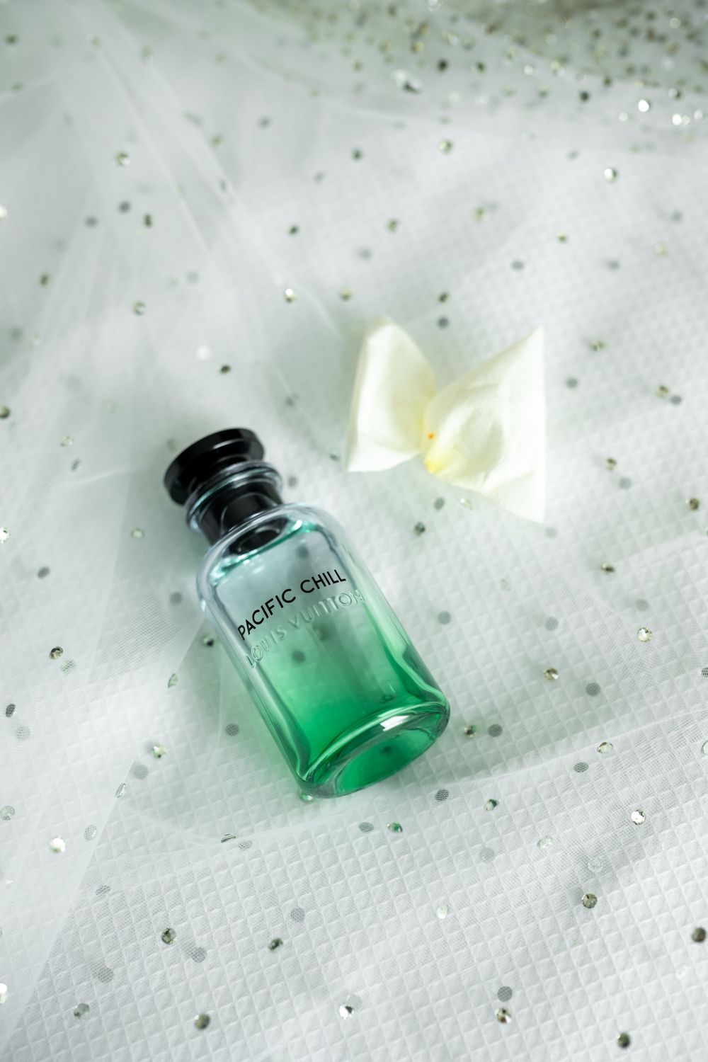 Teal perfume bottle with white flower petals on textured white fabric.