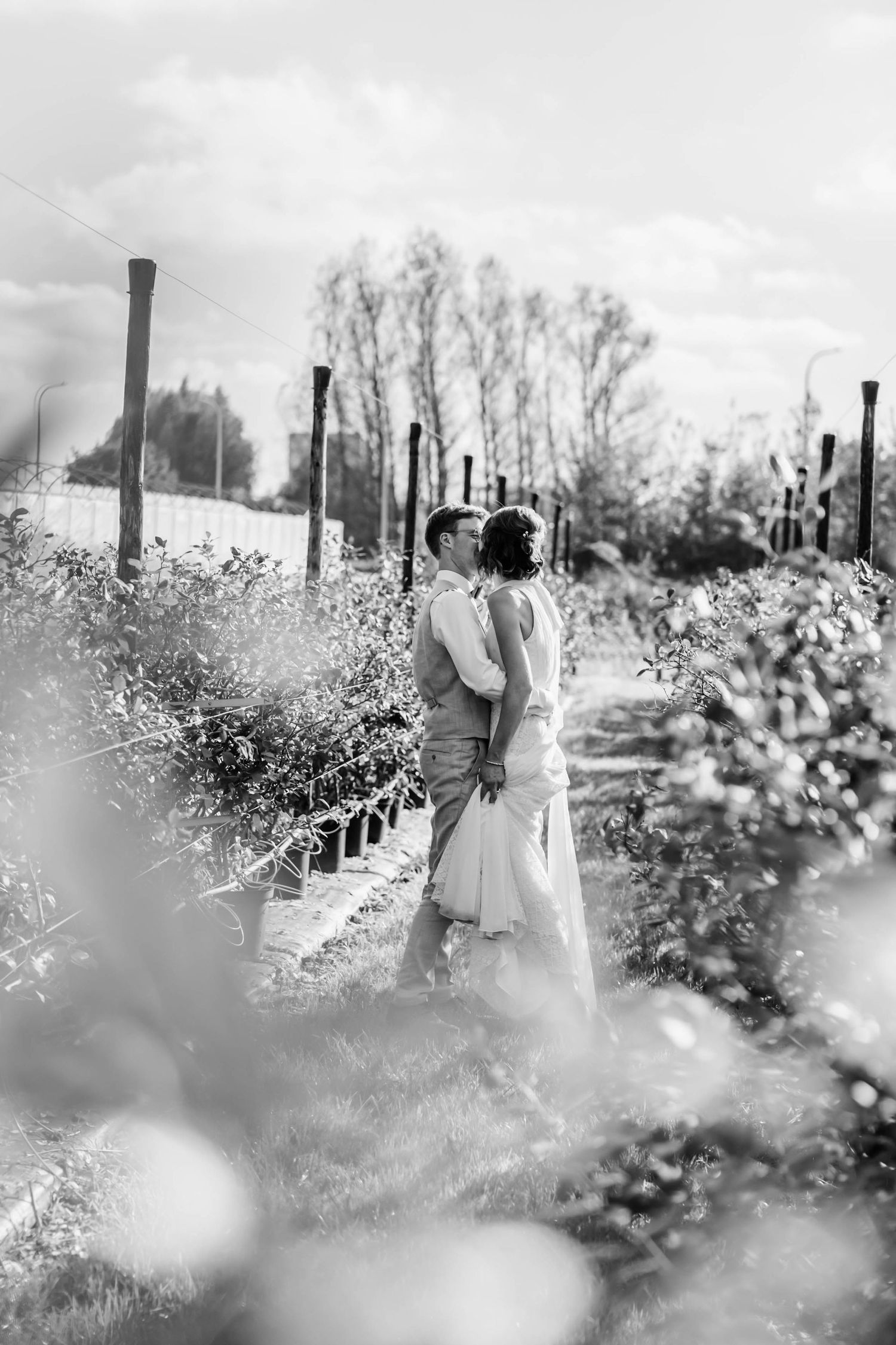 A romantic black and white photo of a couple embracing in a garden with blooming flowers and tall posts in the background.