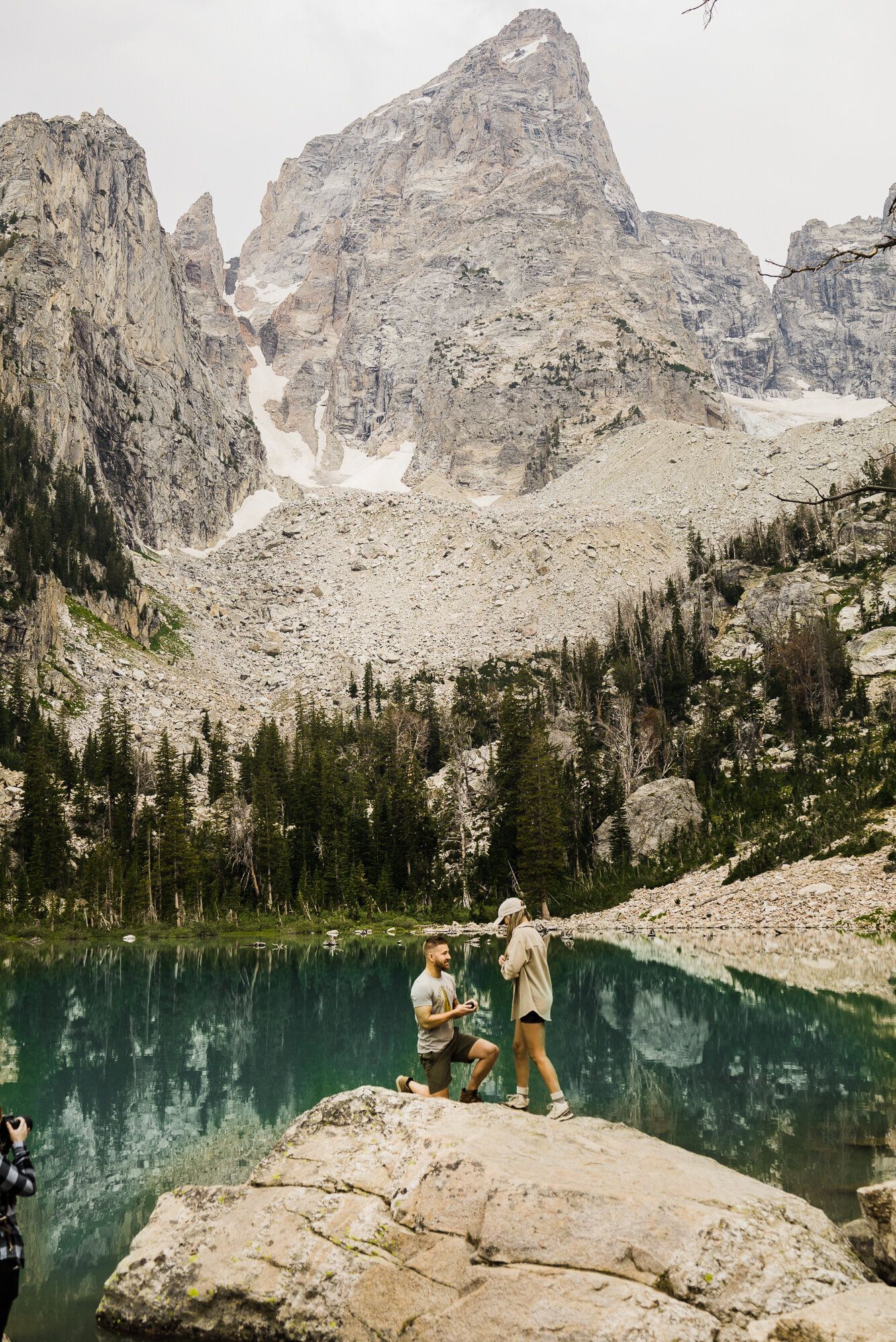Two hikers stand on rocks beside a turquoise alpine lake surrounded by rocky peaks and evergreen forest.
