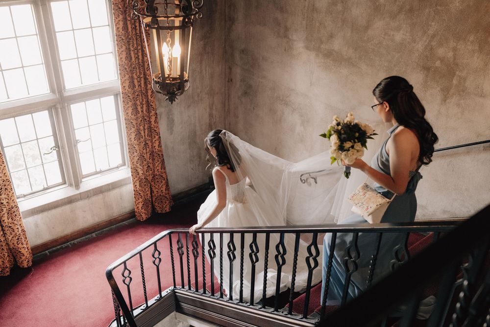 Bridesmaid holding bouquet helps bride with veil on elegant staircase.