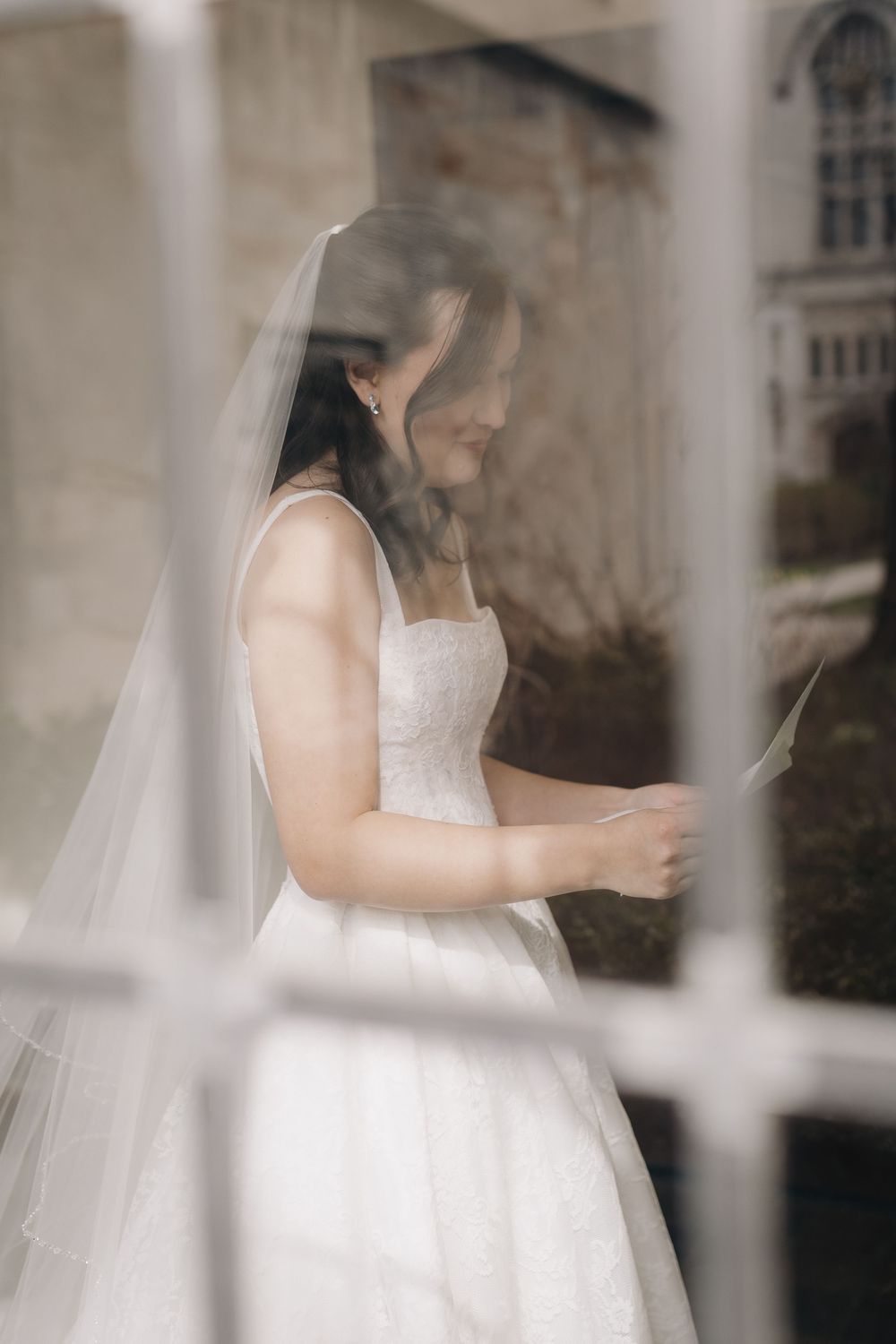 Bride in white gown and veil reading a letter by a window.