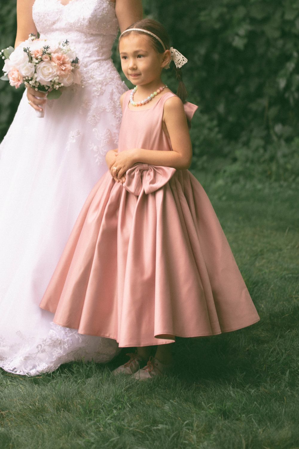 Young flower girl in a blush pink ball gown dress stands beside the bride at an outdoor wedding.