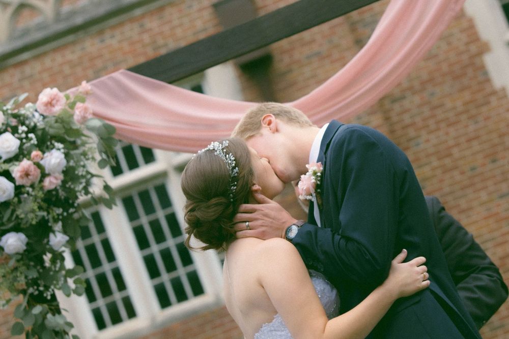 Bride and groom share their first kiss at an outdoor wedding ceremony with pink draping and floral arrangements.