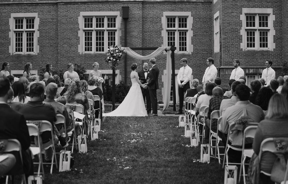 Black and white photo of an outdoor wedding ceremony with guests seated in white chairs before a brick building.