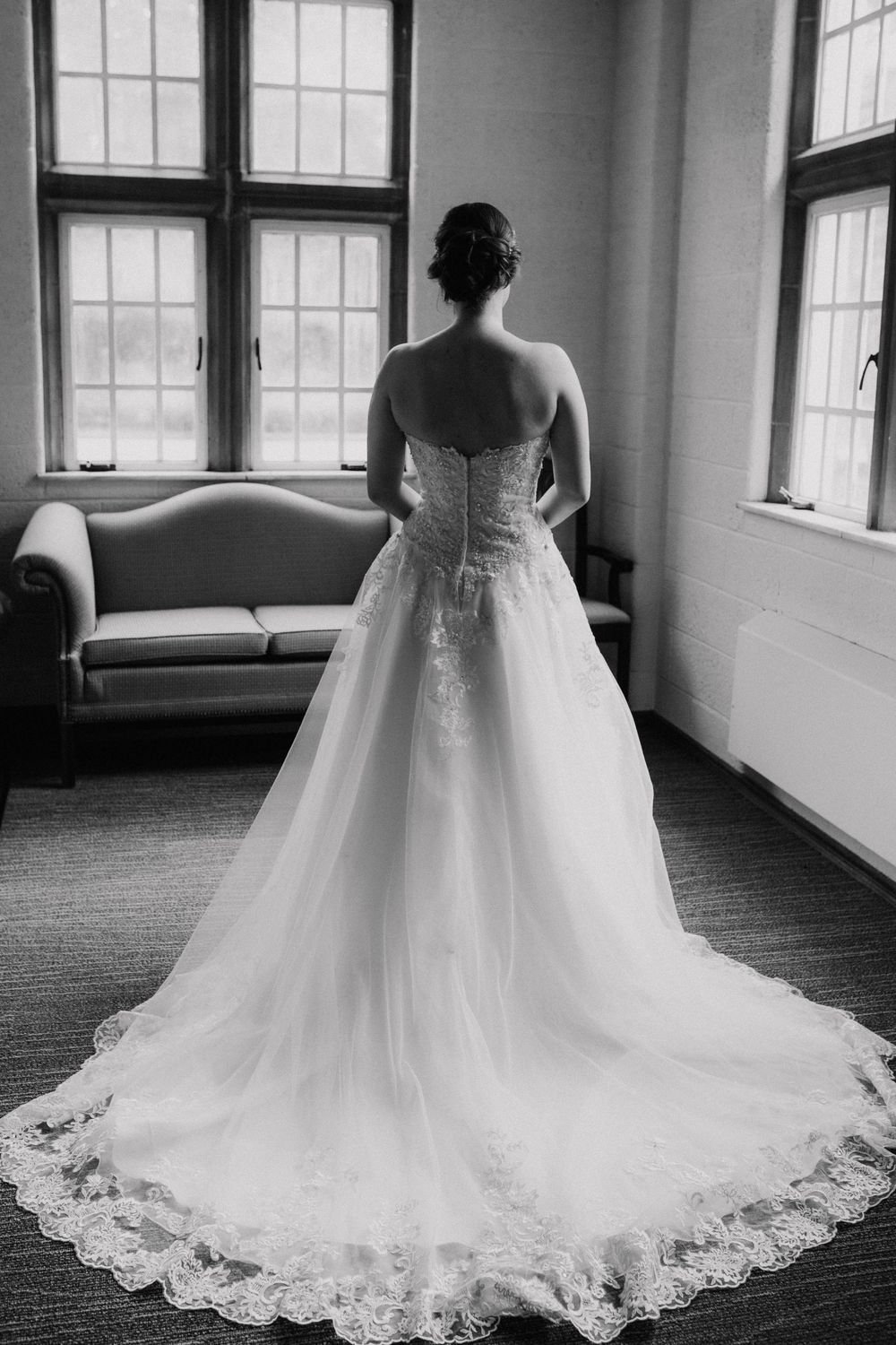 Black and white photo of a bride in a strapless lace ball gown with a long train standing by windows.