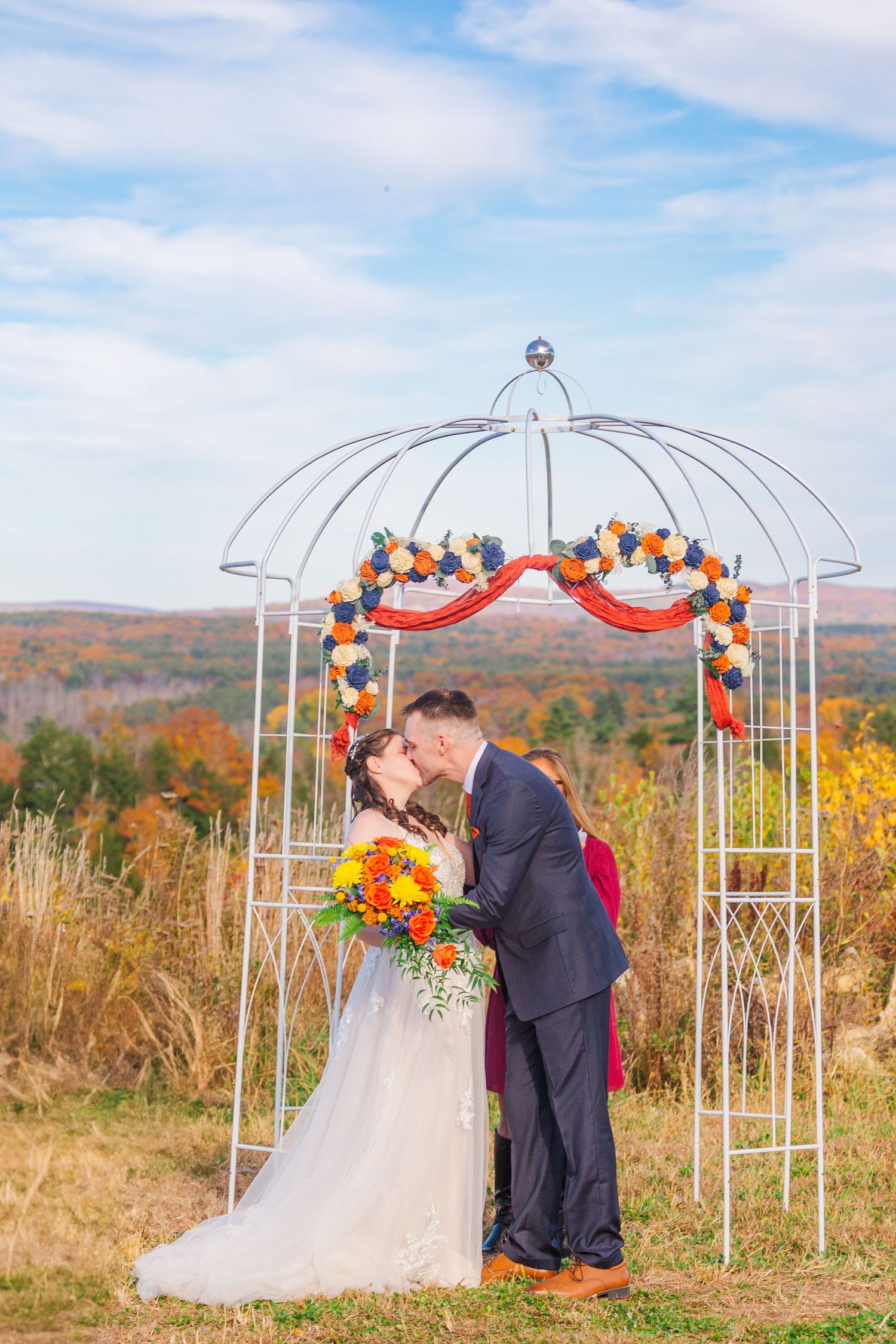 Couple sharing romantic kiss under white metal gazebo decorated with orange and red flowers during autumn wedding.