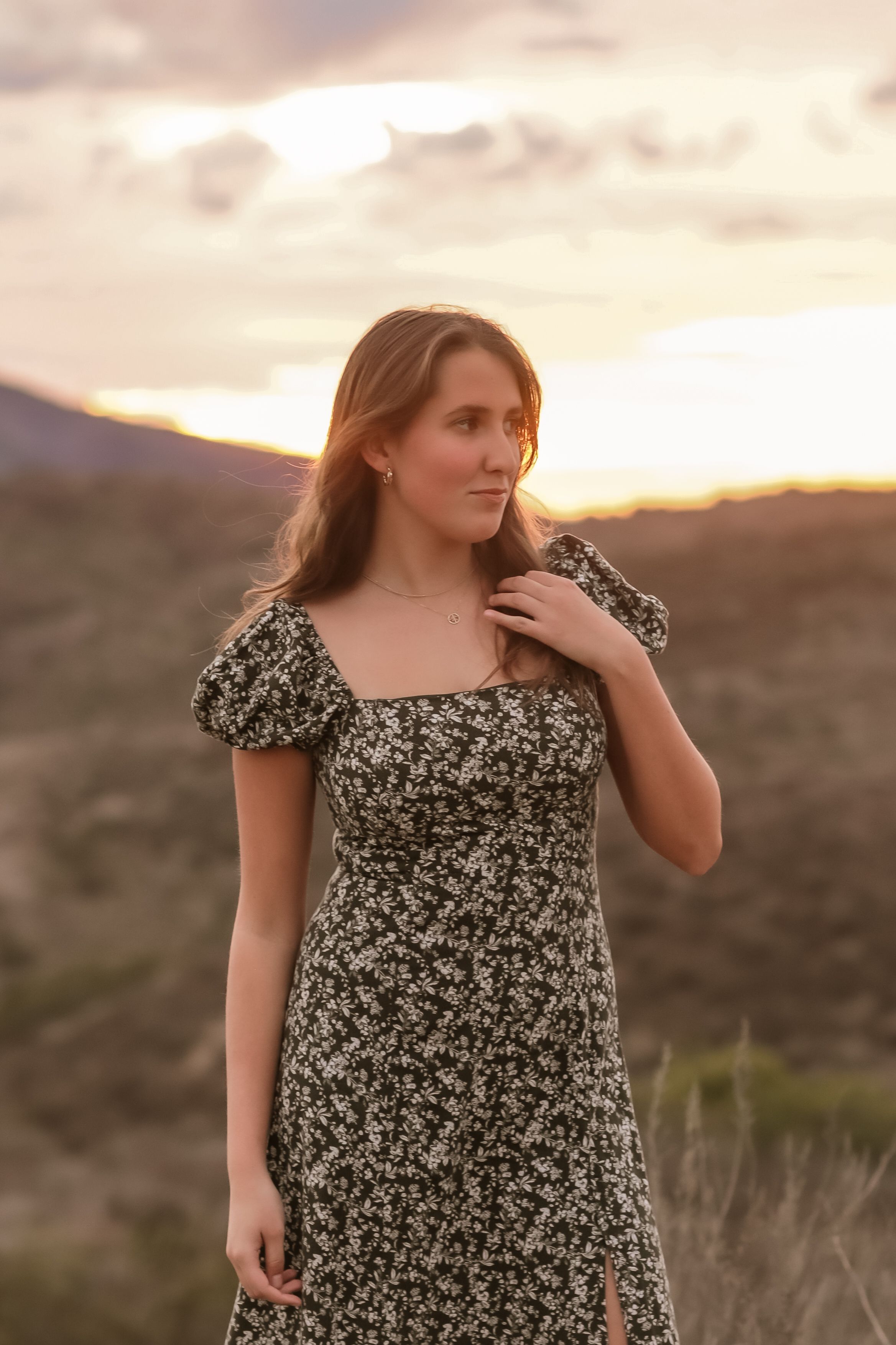 A young person in a floral dress poses at sunset against a mountainous landscape with golden light.
