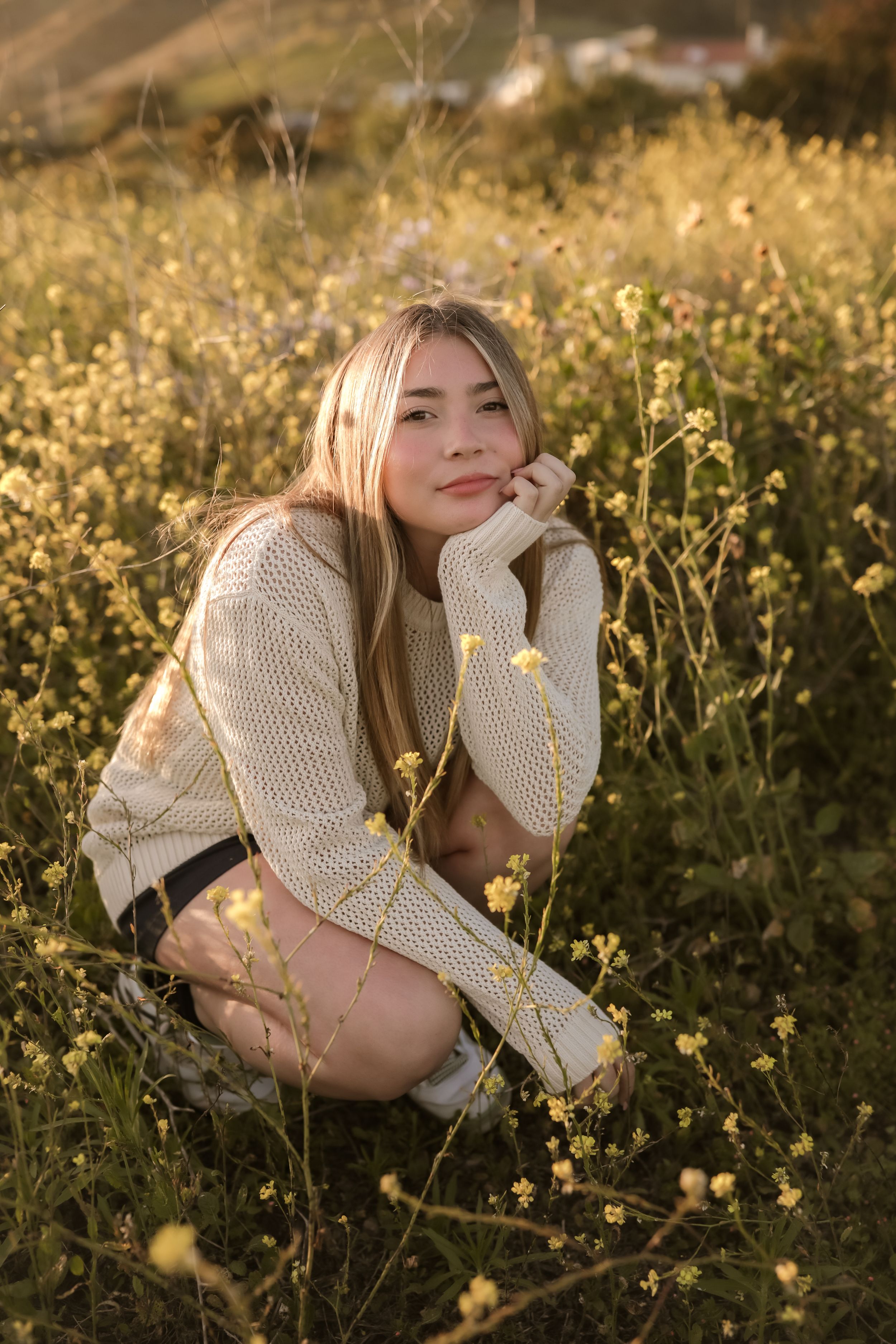 Person in cozy cream sweater crouches among yellow wildflowers in a golden sunlit field during sunset.
