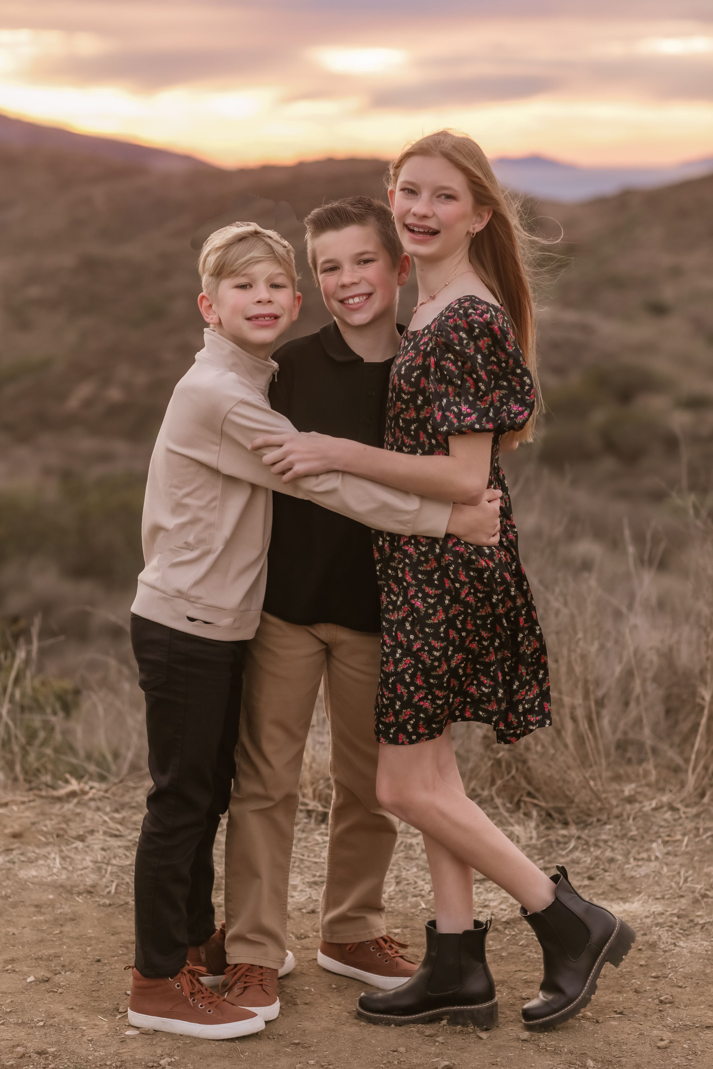 Three siblings share a joyful embrace while posing together on a desert trail at sunset.