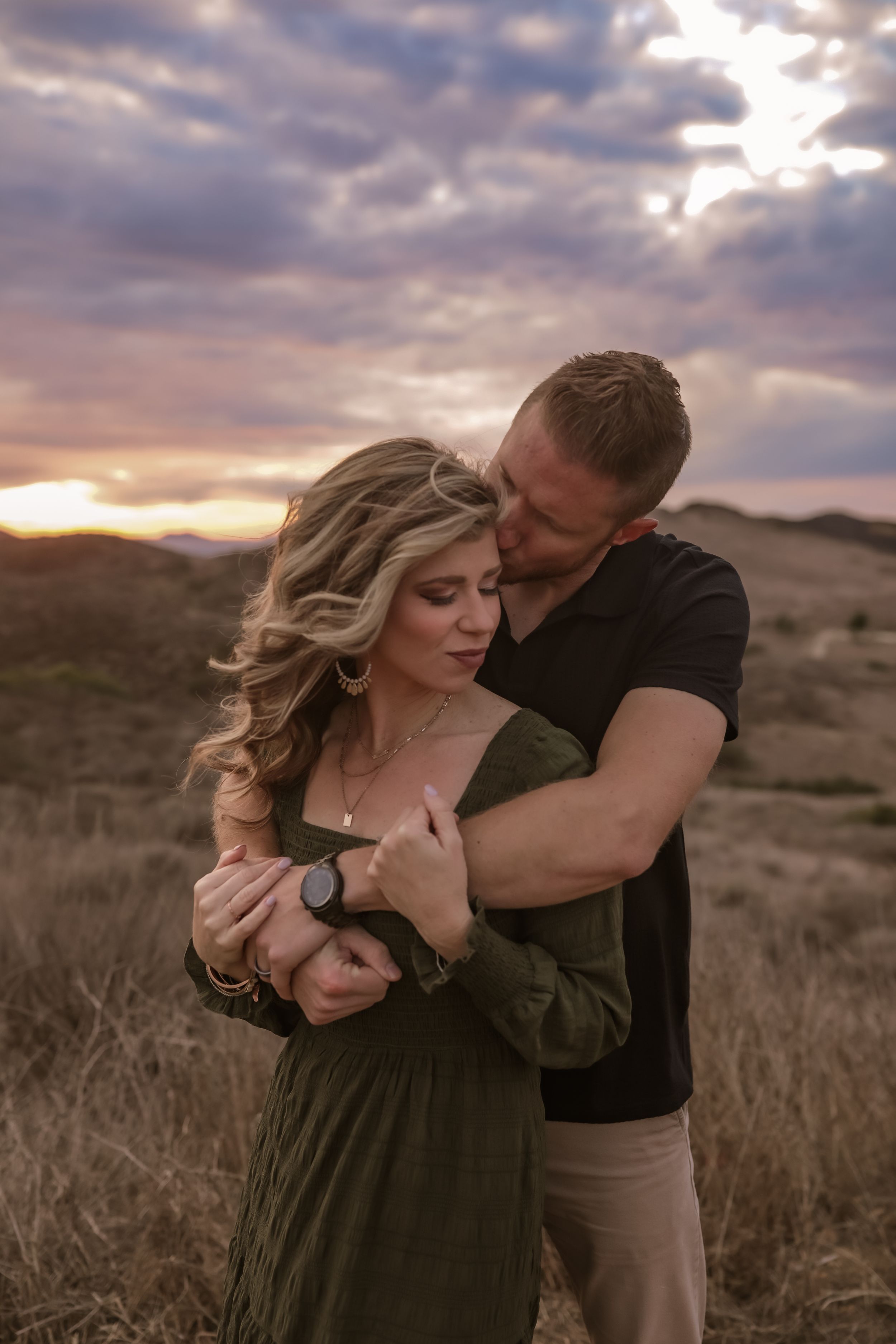 A romantic couple embraces in a desert landscape at sunset with dramatic cloudy skies overhead.
