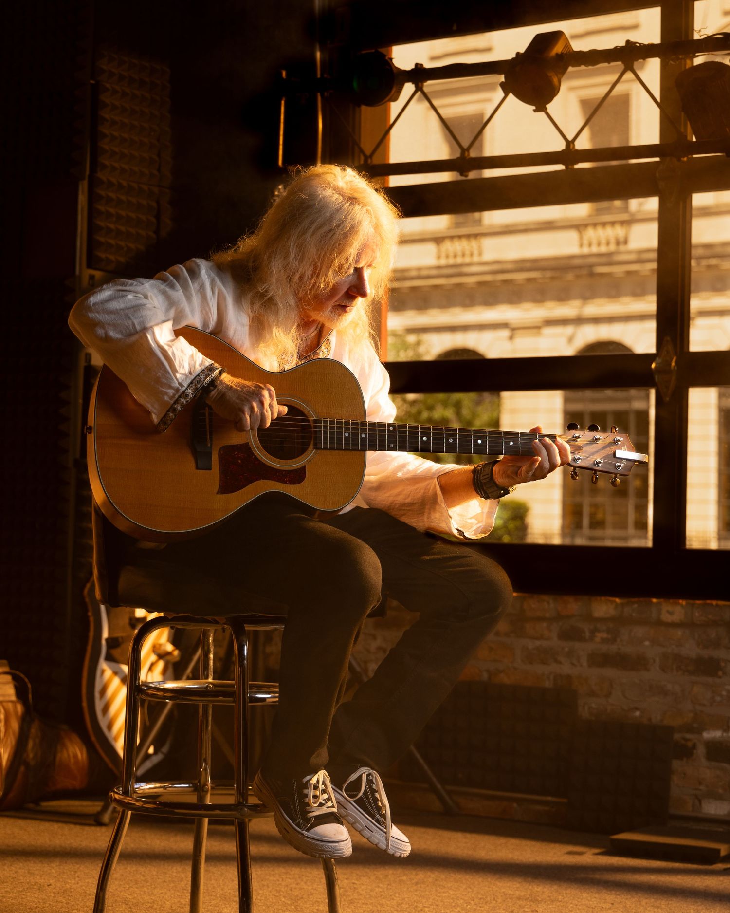 A musician plays acoustic guitar while sitting on a stool by a sunlit window.