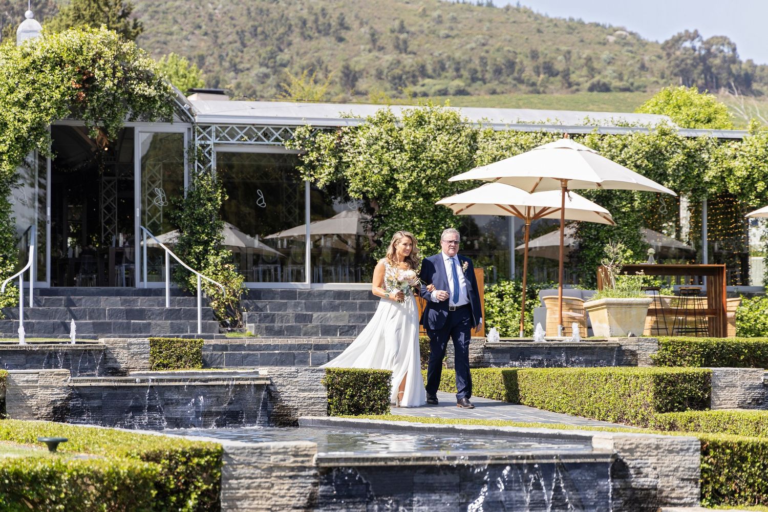 Newlyweds pose on beautiful terraced garden steps with umbrellas and greenery.