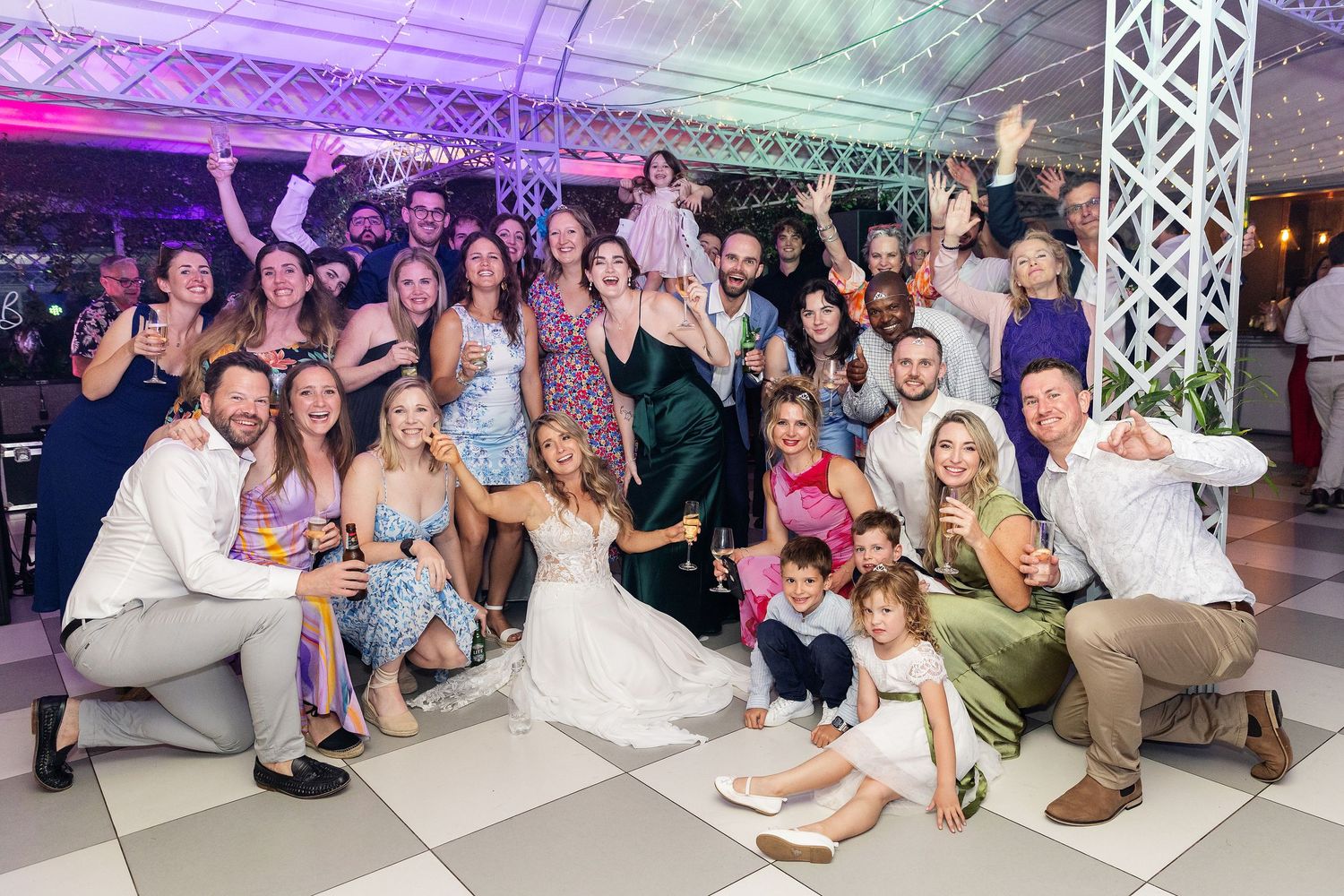 Large wedding party group poses together during reception celebration under purple lighting.