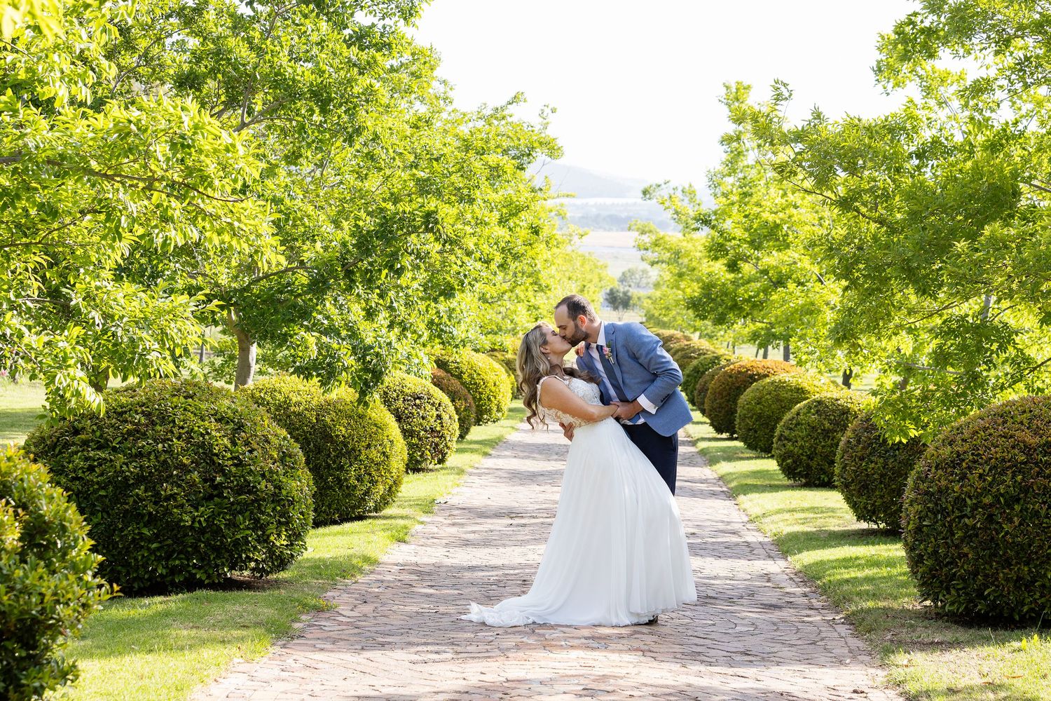 Romantic wedding couple kisses on a garden path lined with manicured shrubs.
