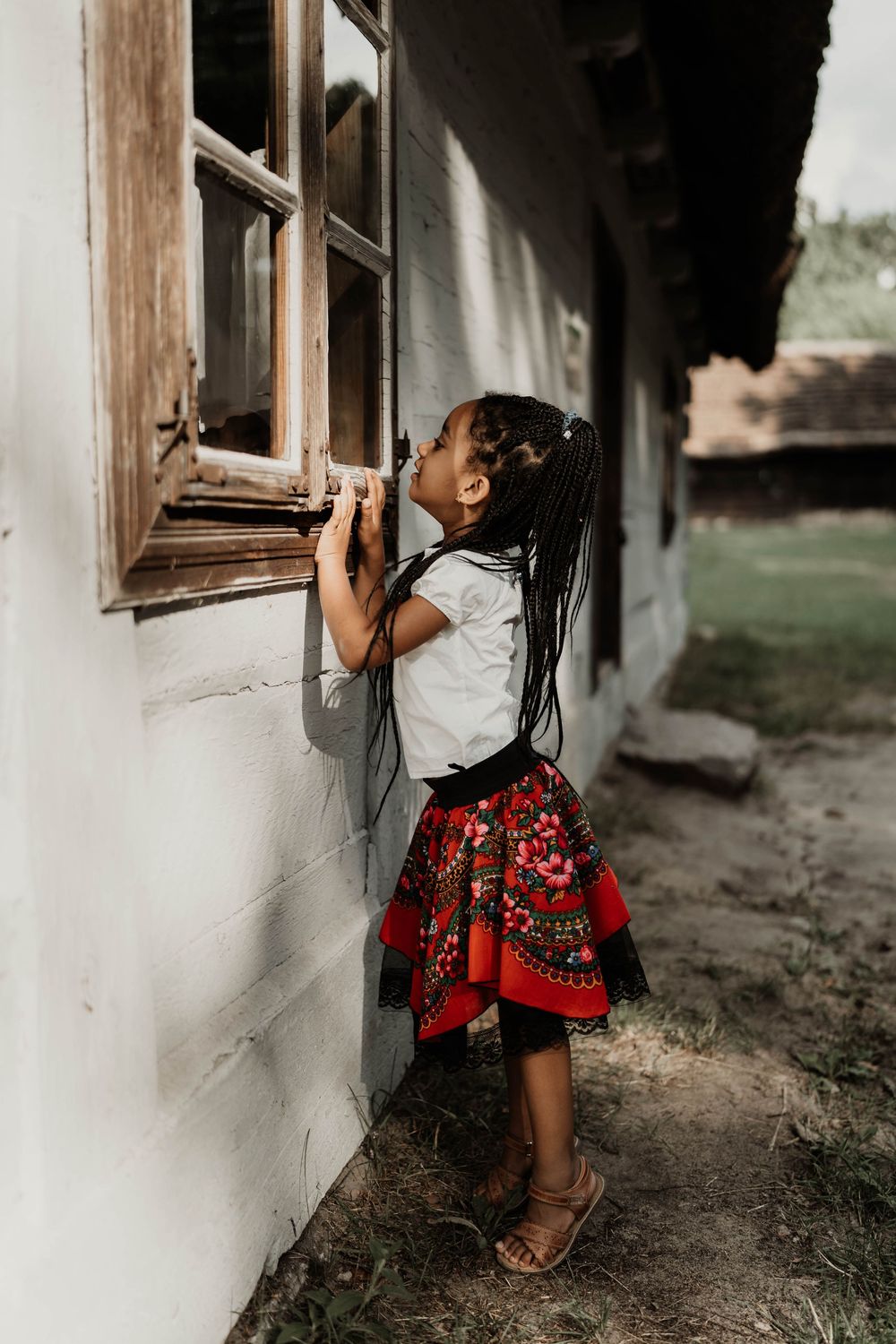 A child in traditional dress peeks through an old wooden window of a rustic building.