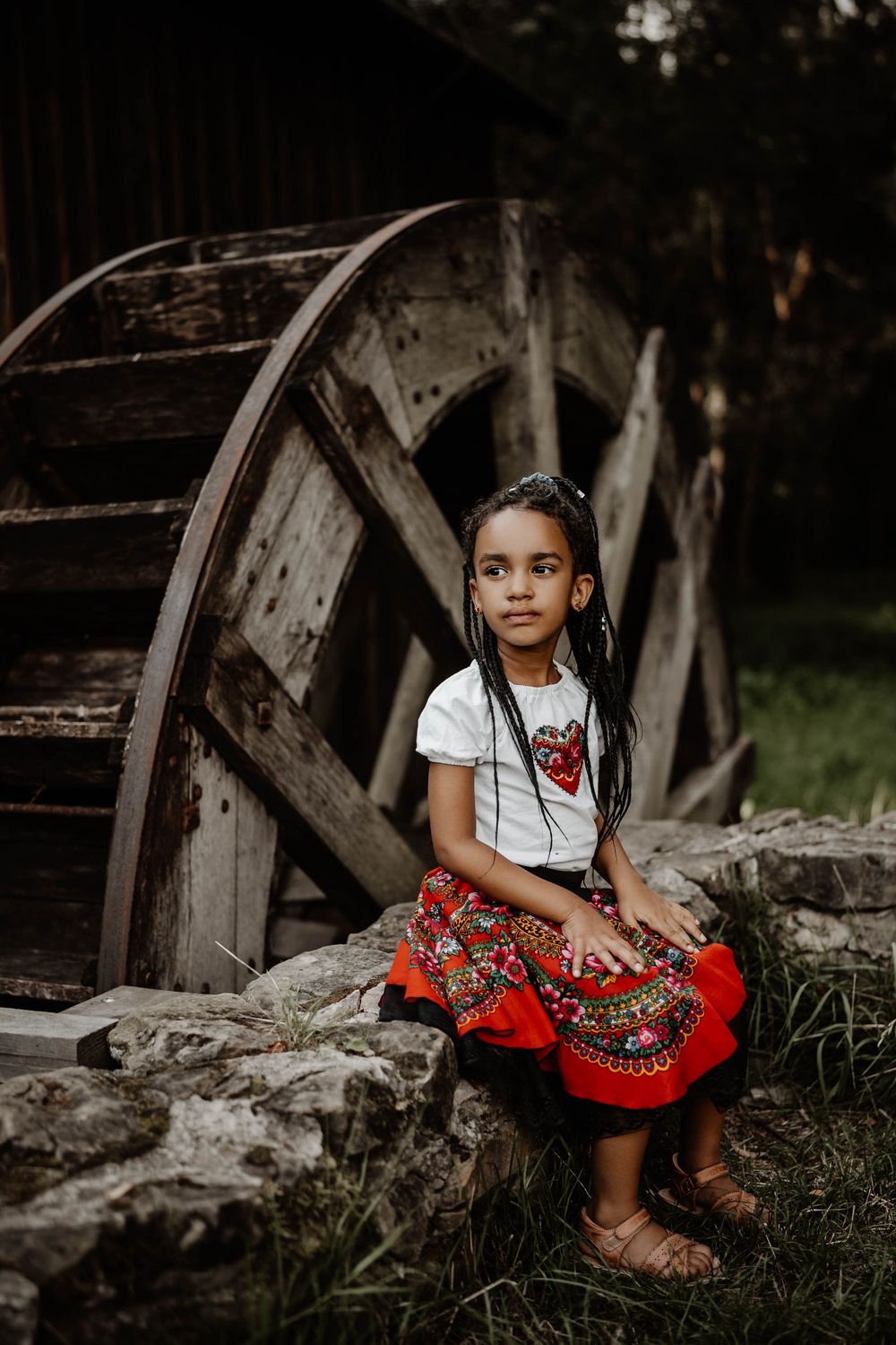 A child in folk dress sits beside an old wooden water wheel at a rustic mill.