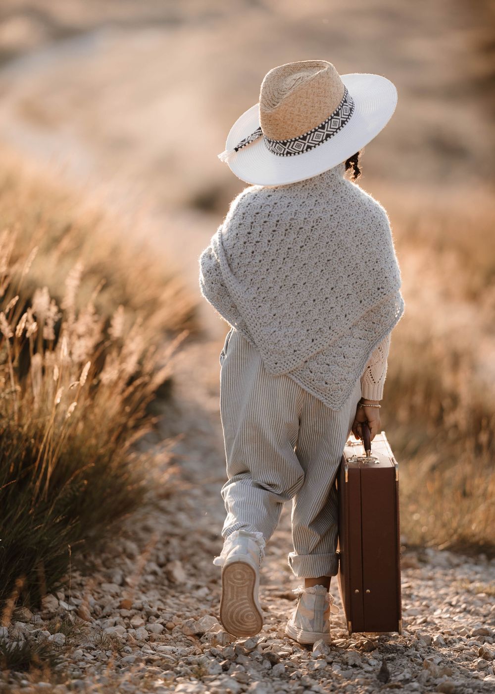 A figure in white wearing a sun hat walks along a path carrying a vintage suitcase.