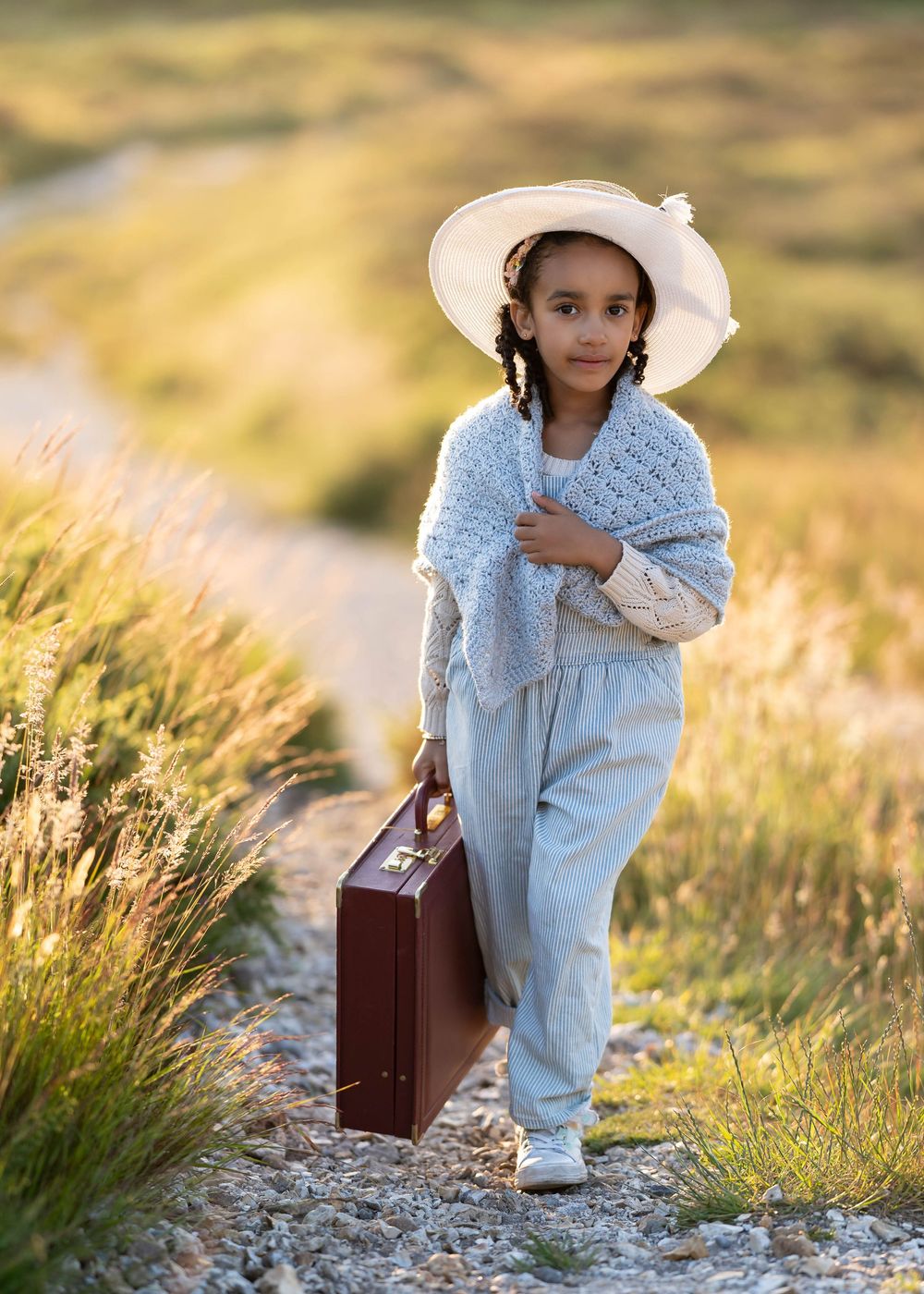 A child in a white sunhat and blue shawl walks along a countryside path carrying a vintage brown suitcase.