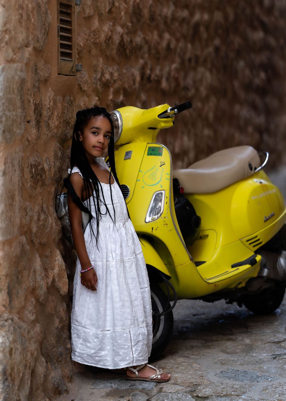 A girl in a white dress standing next to a bright yellow Vespa scooter against a stone wall.