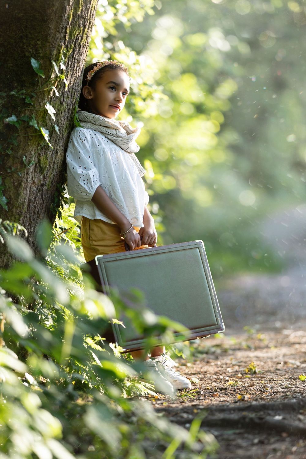 A child in vintage clothing stands by a tree holding a gray suitcase in dappled sunlight.