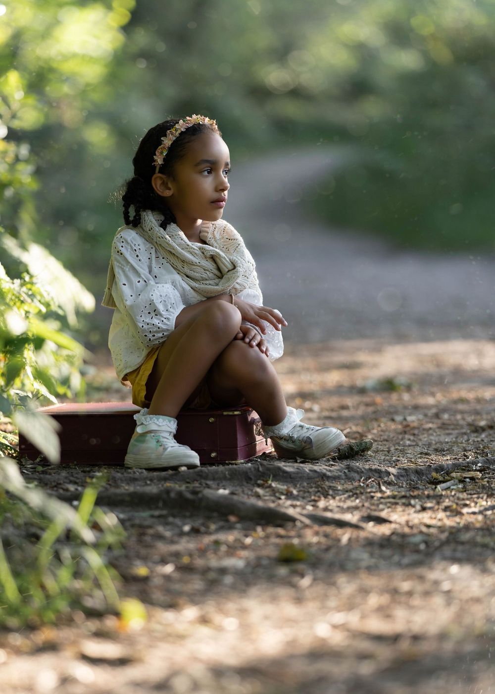 A child in a white sweater and yellow shorts sits on a suitcase along a forest path.