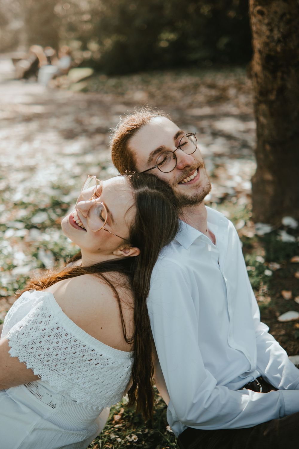 A smiling couple sits outdoors, woman in white off-shoulder top leans back on man wearing glasses and white shirt.