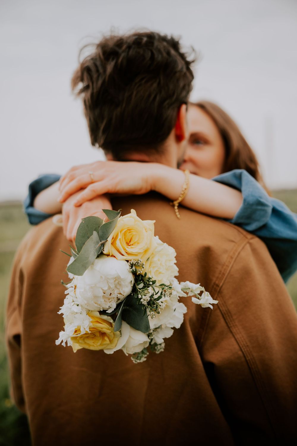 Couple embracing outdoors, woman holding a bouquet of yellow and white roses while hugging man in brown jacket.
