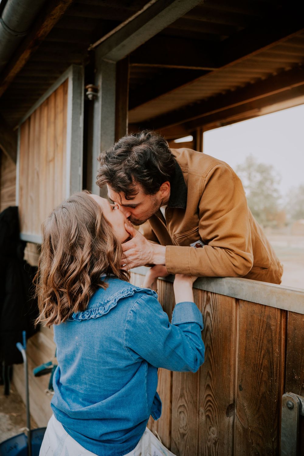 A couple shares a romantic kiss over a wooden fence at a rustic outdoor venue, woman in blue denim jacket.