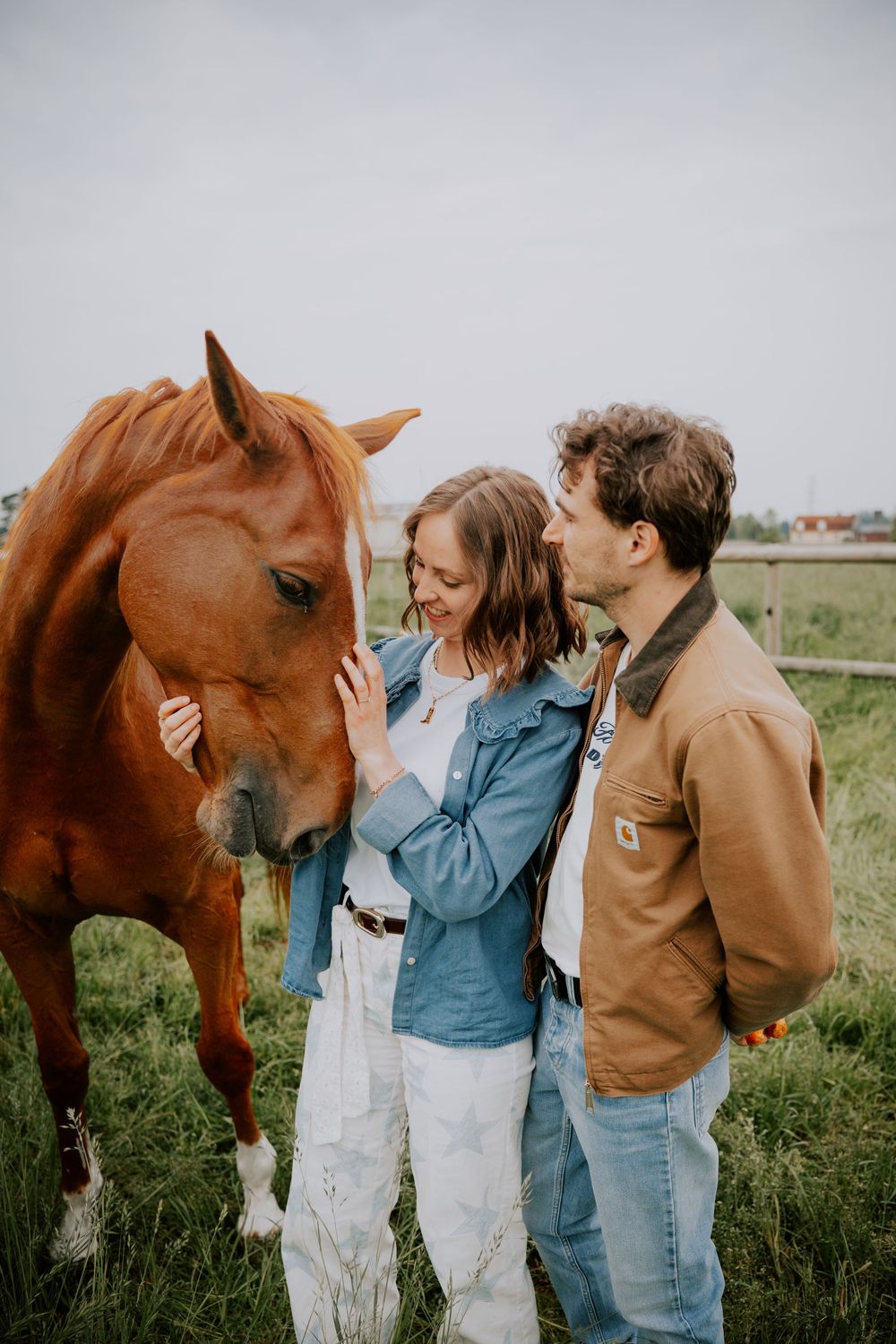 A couple in casual denim outfits tenderly petting a chestnut horse in a green field.